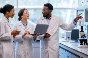 Three scientists in lab coats discuss an experiment: one holds a laptop while another shows notes and a third points to a sample bottle on the bench.