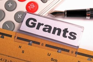 Close-up of a 'Grants' sign resting on a ruler, with a fountain pen and calculator keys in the background to suggest funding and administration.