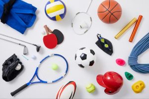 Assorted sports gear scattered on a white surface: soccer ball, volleyball, basketball, tennis racket, jump rope, goggles, whistle, and other accessories.