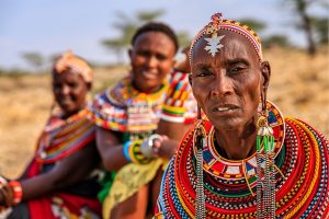 Close-up of a Maasai woman wearing intricate multicolored beadwork and a headpiece, with two other beaded women in a dry landscape in the background.