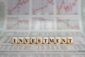Wooden letter blocks spell INVESTMENT on a newspaper with blurred stock charts in the background.