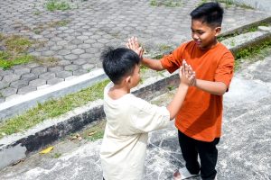 Two boys high-fiving each other outdoors on a sunny day by a paved walkway.