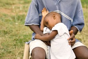 Adult in a blue shirt sits outdoors with a toddler resting asleep on their lap and head on the arm.