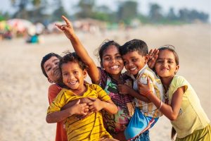 Five smiling children hug each other on a sandy beach, enjoying a sunny day together.
