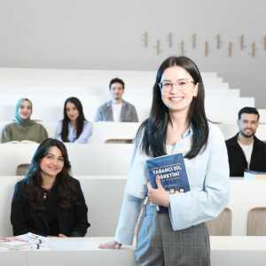 Smiling young woman with glasses standing in a bright classroom, holding a blue book titled Yabancı Dil Öğretimi.