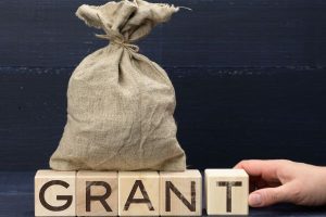 Burlap money bag on top of wooden blocks spelling GRANT, with a hand to the right adjusting them.