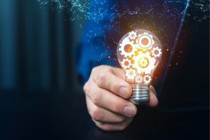 Hand gripping a lit light bulb containing gears, symbolizing innovation and engineering ideas.