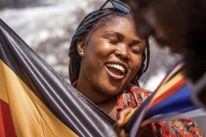 Joyful woman with braided hair and sunglasses on her head, smiling while holding a colorful flag outdoors.