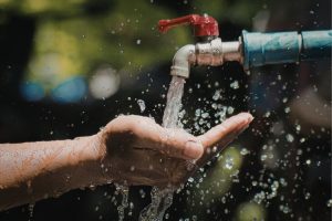 Hands held under a stream of water from an outdoor faucet with a red handle, water droplets splashing around.