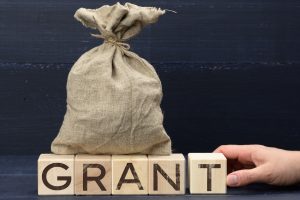 Burlap sack sits on wooden blocks spelling GRANT, with a hand resting nearby, symbolizing a grant.