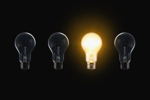 Four light bulbs in a row on a black background; the third bulb is lit and glowing yellow from the center-left position from the left of the row.