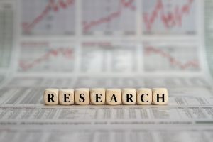 Wooden letter blocks spell RESEARCH on a newspaper, with stock charts blurred in the background.