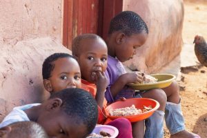 Three young children sit against a dusty wall eating from colorful plastic bowls outside, with a chicken nearby.