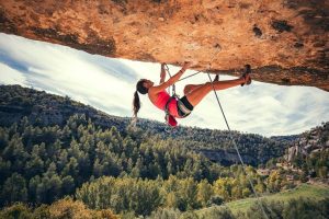 Female climber in a red top ascending an overhanging rock face with a rope, forested valley in the background.