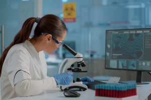 Female scientist in a lab coat, safety goggles, and blue gloves uses a microscope beside a computer monitor in a modern lab.