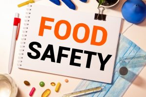Notebook page reads FOOD SAFETY in bold orange and black, surrounded by gloves, a mask, and PPE on a desk.