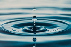 Close-up of a water droplet about to hit the blue surface, forming circular ripples on calm water.