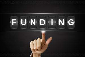 A hand pointing at the word FUNDING displayed as flipping tiles, representing selecting funding options or access to funds.