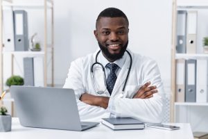 Friendly Black male doctor in a white coat with a stethoscope sits at a desk, arms crossed, smiling at the camera with a laptop and notebook nearby.