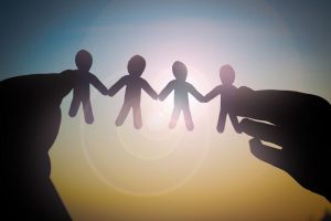 Silhouetted hands hold a chain of paper dolls linked by the hands against a sunset sky, symbolizing unity and teamwork.