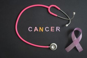 Pink stethoscope loop with the word CANCER in colorful letters and a purple awareness ribbon on a dark background, symbolizing cancer awareness.