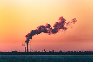 Industrial smokestacks releasing dark smoke along a coastline at sunset.