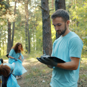 Volunteer in a teal shirt holds a clipboard while picking up litter in a forest. (Informative)