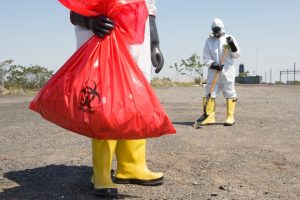 Two hazmat-suited workers clean a barren lot; foreground worker holds a large red biohazard bag, background worker with a shovel.