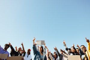Diverse crowd of protesters holding up signs and raising their fists against a clear blue sky.