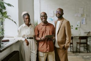 Three professionals posing together in a bright office space, smiling at the camera; the middle person holds a tablet.